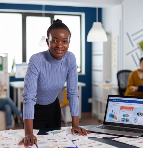 Dark skinned woman and colleagues in corporate start up office working to finish project. Diverse team of business people analyzing company financial reports from computer.
